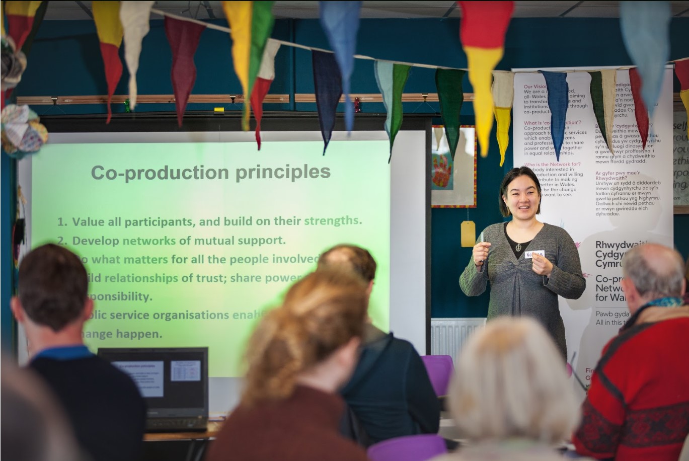 Noreen is presenting in front of slides titled 'Co-production principles' to a room of people