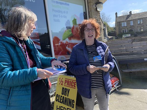 Two women with clipboards and 'Ask Shepley volunteer' badges outside a co-op